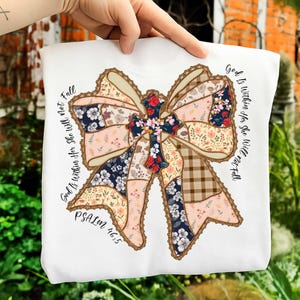 May include: White sweatshirt featuring a large, patchwork bow design with floral and plaid patterns. The bow has a cross in the center and the text "God Is Within Her She Will Not Fall" and "PSALM 46:5" in black script.