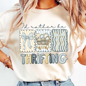 May include: Cream-colored t-shirt with the text "I'd rather be thrifting." The design features three rectangular panels with a bow, a shopping cart filled with dishes, and another bow. The word "THRIFTING" is in large letters.
