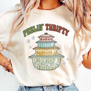May include: Cream-colored t-shirt featuring a stack of colorful, vintage-style casserole dishes. The text "FEELIN' THRIFTY" arches above the dishes in green and red lettering. Daisy accents are scattered around the design.