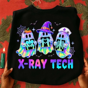 May include: A black sweatshirt featuring a graphic of three iridescent ghosts holding X-ray images. The ghosts wear a pumpkin, a witch hat, and a nurse cap. The text X-RAY TECH is printed in a gradient of purple to blue below the ghosts.