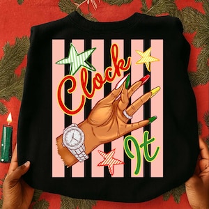 Puede incluir: Sudadera negra con un gráfico de una mano con reloj y anillos, con uñas largas y coloridas. Las palabras "Clock It" están escritas en escritura roja y verde, con acentos de estrellas y rayas rosas y negras.