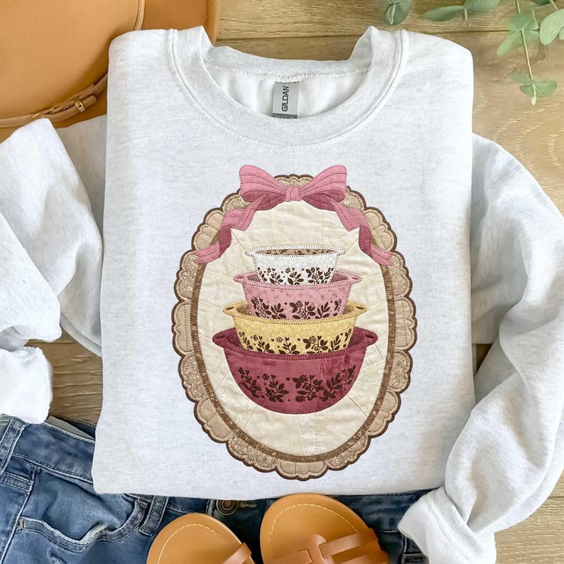 May include: A light gray sweatshirt featuring a decorative oval design with a stack of four floral patterned mixing bowls. A pink bow is at the top of the oval. The bowls are in shades of white, pink, yellow, and burgundy.