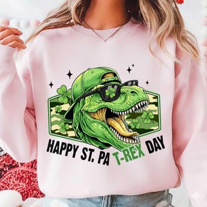 May include: Pale pink sweatshirt with a green dinosaur wearing sunglasses and a cap, with the text "HAPPY ST. PA T-REX DAY". The dinosaur graphic includes shamrocks and a camouflage background, ideal for St. Patrick's Day.