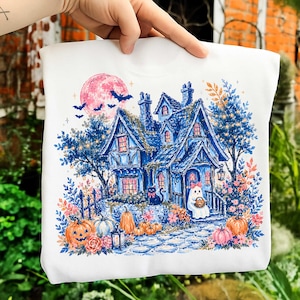 Pu&ograve; includere: Una felpa bianca con un'illustrazione dettagliata di una casa blu, una luna rosa, pipistrelli, un gatto nero e un fantasma che tiene un cestino. La scena include zucche arancioni e bianche, fiori colorati e un sentiero in pietra in giardino.
