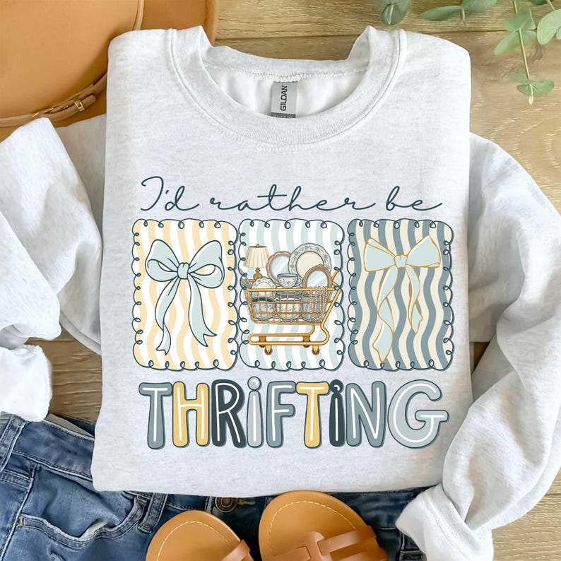 May include: Light gray sweatshirt with the text "I'd rather be THRIFTING." The design includes three framed illustrations: a bow, a shopping cart filled with dishes, and a striped pattern with a bow. The sweatshirt is paired with blue jeans and sandals.