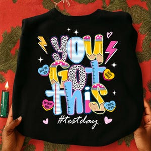 May include: Black sweatshirt featuring the phrase "YOU GOT THIS" in a vibrant, cartoonish style. The design incorporates pencils, hearts, and lightning bolts, with the hashtag "#testday". A cheerful and encouraging design for exam day.