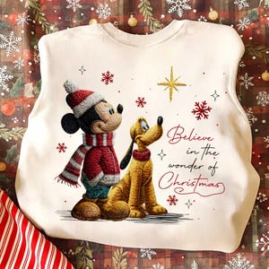 May include: Cream-colored sweatshirt featuring Mickey Mouse and Pluto in winter attire. Mickey wears a red hat and scarf, Pluto a red collar. The text "Believe in the wonder of Christmas" is written in red script, with snowflakes and a star.