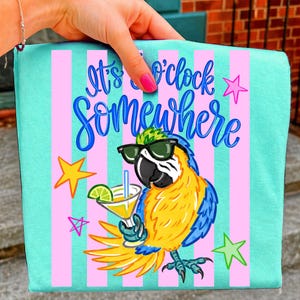 Puede incluir: Un bolso tote turquesa con rayas verticales rosas y la frase "It's o'clock somewhere" en escritura azul. Se presenta un loro de dibujos animados con gafas de sol y sosteniendo un cóctel. También se incluyen estrellas amarillas y rosas.