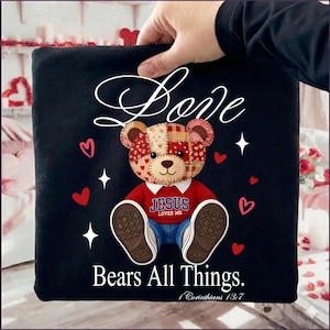 May include: Black square pillow with a teddy bear design. The bear has a patchwork head, a red shirt with "JESUS LOVES ME", and blue jeans. The pillow also has the word "Love" and "Bears All Things" with hearts and stars.