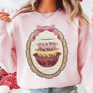 May include: A light pink sweatshirt featuring a decorative oval design with a stack of four floral patterned mixing bowls in shades of white, pink, and yellow. A pink bow and ribbon are at the top of the oval.