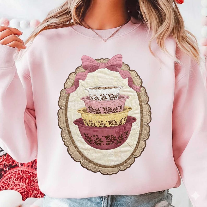 May include: A light pink sweatshirt featuring a decorative oval design with a stack of four floral patterned mixing bowls in shades of white, pink, and yellow. A pink bow and ribbon are at the top of the oval.