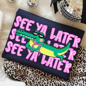 May include: Navy blue sweatshirt with a graphic of a green alligator wearing sunglasses and a pink bow tie. The text "SEE YA LATER" is in pink, outlined in white. The alligator has a yellow checkered belly.