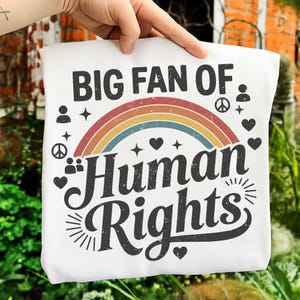 May include: White tote bag with the text "BIG FAN OF HUMAN RIGHTS" in black, arched over a rainbow. The design includes hearts, stars, and peace symbols. The bag is held up by a hand.