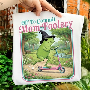 Puede incluir: Bolso tote blanco con una rana verde con un sombrero de bruja negro, montada en un patinete rosa y azul. El bolso tiene el texto "Off To Commit Mom-Foolery" en rosa y turquesa, con un fondo de escena de parque.