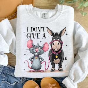 Puede incluir: Una sudadera gris claro con un rat&oacute;n y un burro de dibujos animados y el texto "I DON'T GIVE A." El rat&oacute;n es gris con orejas rosas y un collar de estrella. El burro es negro con un cabestro rosa.