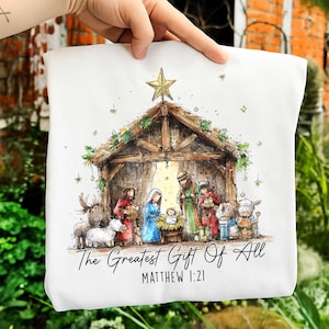May include: White fabric with a watercolor illustration of the Nativity scene. The image features a wooden stable, a golden star, and figures of Mary, Joseph, and baby Jesus. Text reads "The Greatest Gift Of All" and "Matthew 1:21".