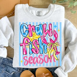 May include: A light gray sweatshirt with a colorful graphic that says "Crawfish Season." The graphic features a pink crawfish with a yellow bow, set against a blue and white striped background. The text is in blue and pink.