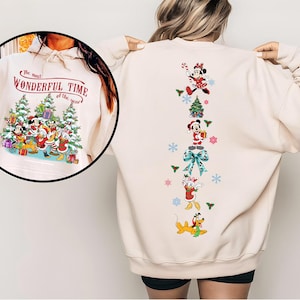 May include: Cream-colored sweatshirt featuring a Christmas-themed design. The front displays a graphic with Mickey Mouse, Donald Duck, and other characters with the text "The most Wonderful Time of the Year." The back has a vertical arrangement of Christmas characters and decorations.