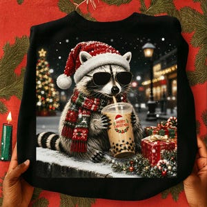 May include: A black sweatshirt featuring a graphic of a raccoon wearing a red Santa hat and black sunglasses, wrapped in a red and green patterned scarf. The raccoon holds a plastic cup with boba tea, labeled with the text Merry Christmas.