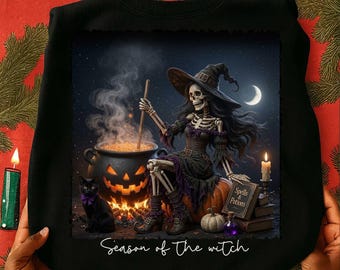 Op de afbeelding: Een zwart sweatshirt met een afbeelding van een skeletheks met een hoed zittend op een pompoen, roerend in een gloeiende pompoenketel met een zwarte kat ernaast. Het beeld bevat een boek met de titel Spells & Potions en de tekst Season of the witch.