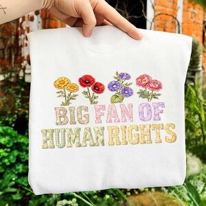 Könnte beinhalten: Ein weißes Sweatshirt mit bunten Blumenstickereien und dem Schriftzug "BIG FAN OF HUMAN RIGHTS" in einer Mischung aus gemusterten Schriftarten. Das Design umfasst gelbe, rote, violette und rosa Blumen.