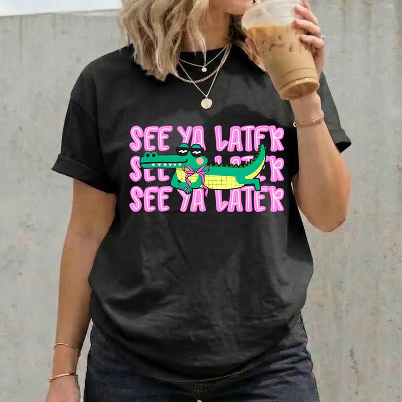 May include: Black t-shirt with a graphic of a green alligator wearing a bow tie, and the text "SEE YA LATER" in pink. The alligator is smiling. The shirt is a casual style.