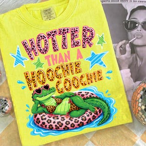 May include: Yellow t-shirt with the text "HOTTER THAN A HOOCHIE COOCHIE" in pink leopard print lettering. The design includes a cartoon alligator wearing sunglasses and a bikini, lounging on a leopard print inner tube. Blue and green stars and water droplets complete the design.