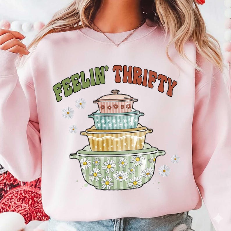 May include: A light pink sweatshirt with the words "FEELIN' THRIFTY" in green and orange. The graphic features a stack of vintage-style casserole dishes in various colors and patterns, with daisy accents.