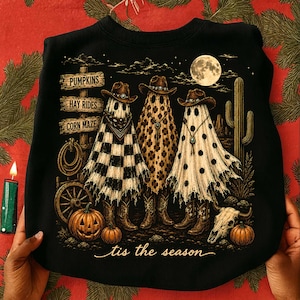 May include: A black sweatshirt featuring a graphic of three ghosts wearing cowboy hats and boots. The ghosts have checkered, leopard, and polka dot patterns. The design includes a signpost with text PUMPKINS, HAY RIDES, CORN MAZE, and the text 'tis the season.