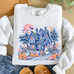 Pu&ograve; includere: Una felpa girocollo grigio chiaro con una stampa grafica di una casa blu con una luna rosa, pipistrelli, un gatto nero e un fantasma. Il design include zucche arancioni, fogliame autunnale e un sentiero in pietra verso la porta d'ingresso.