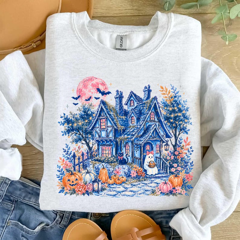 Pu&ograve; includere: Una felpa girocollo grigio chiaro con una stampa grafica di una casa blu con una luna rosa, pipistrelli, un gatto nero e un fantasma. Il design include zucche arancioni, fogliame autunnale e un sentiero in pietra verso la porta d'ingresso.