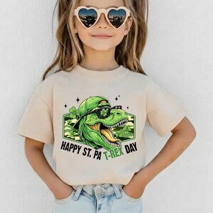May include: A beige t-shirt with a green dinosaur wearing sunglasses and a cap, and the text "Happy St. Pa T-Rex Day". The dinosaur graphic is set against a camouflage background with shamrocks.