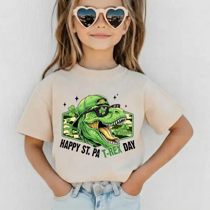 May include: A beige t-shirt with a green dinosaur wearing sunglasses and a cap, and the text "Happy St. Pa T-Rex Day". The dinosaur graphic is set against a camouflage background with shamrocks.