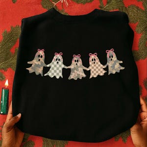 May include: A black crewneck sweatshirt featuring a graphic of five ghosts holding hands in a row. Each ghost has a unique checkered or camouflage pattern in muted tones and wears a small pink bow on its head. The ghosts have black oval eyes.