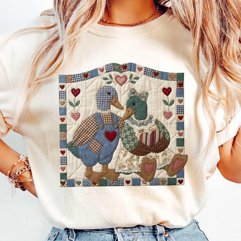 May include: Cream-colored sweatshirt featuring a patchwork design of two ducks in love. The ducks are adorned with heart-shaped accents and dressed in patterned clothing. The design is framed with a quilted border, creating a cozy, country aesthetic.