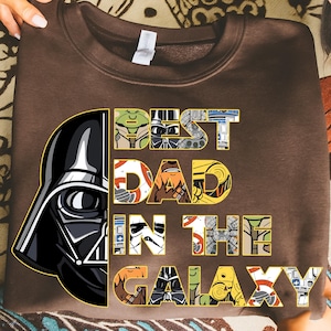 May include: Brown sweatshirt with a graphic of Darth Vader and the text "BEST DAD IN THE GALAXY" in a colorful, Star Wars-themed design. The letters are filled with images of Star Wars characters and elements.