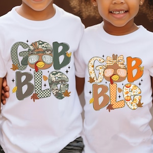 May include: Two white t-shirts with a colorful Thanksgiving-themed graphic. The graphic says "GOBBLE BLG" with a turkey wearing sunglasses and fall leaves. One shirt has a camouflage design, and the other has an orange pumpkin design.