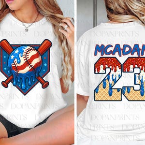 May include: White t-shirt featuring a baseball design with a patriotic theme. The front graphic includes a baseball with red and white stripes and blue stars, with crossed bats. The back displays the name "MCADAM" and the number "23".