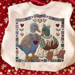 May include: Cream-colored sweatshirt featuring a quilted design of two ducks in love. The male duck wears blue overalls with a red heart, while the female duck has a floral dress and bow. The design includes hearts and a patchwork border.