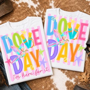May include: Two white t-shirts featuring a colorful graphic with the text DOVE DAY and I'm here for it. The design includes a multi-colored dove, binoculars, and stars over a background of vertical pink stripes. The shirts have a crew neckline.