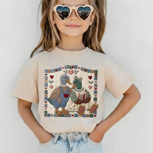 May include: A cream-colored t-shirt featuring a patchwork design of two ducks. One duck wears blue overalls with a red heart, the other a floral dress. The design includes hearts and a quilted border.