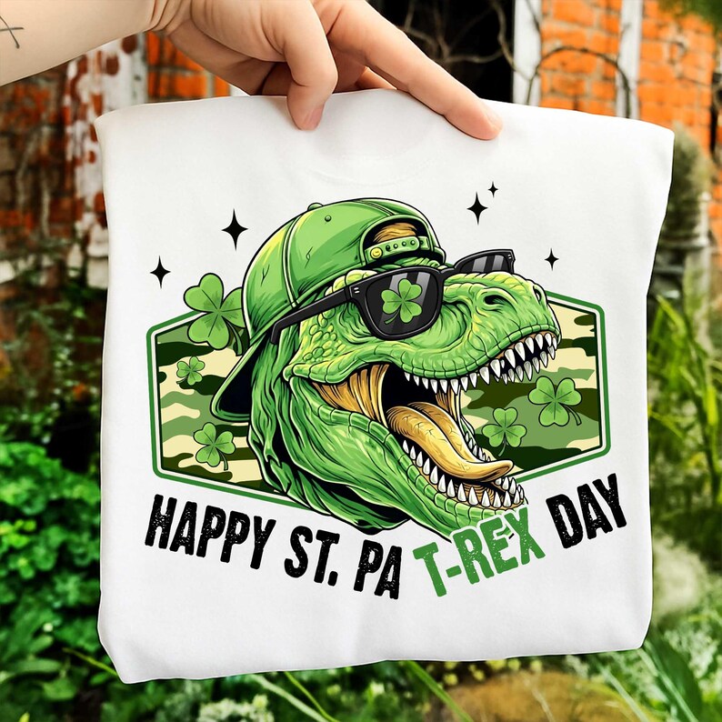 May include: White sweatshirt with a green dinosaur wearing sunglasses and a green cap, with the text "HAPPY ST. PA T-REX DAY". The dinosaur has a green tongue and is surrounded by shamrocks.