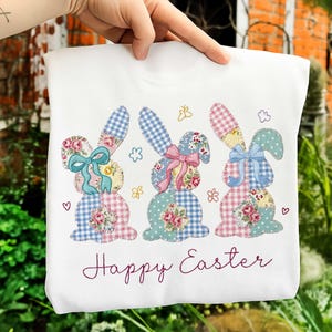May include: White sweatshirt featuring three patchwork bunny rabbits with floral and gingham patterns, each with a bow. The words "Happy Easter" are embroidered in a cursive font below the bunnies. The design includes small hearts and flower details.