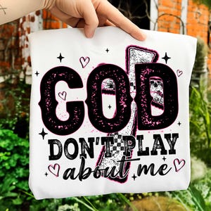 May include: White t-shirt with the text "GOD DON'T PLAY about me" in black, outlined in pink. The design includes hearts and stars. The shirt is held up by a hand.