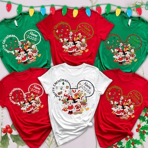 Op de afbeelding: Kerst-thema T-shirts in rood, groen en wit. Elk shirt heeft een cartoon karakter ontwerp met de tekst "Merry Christmas" en "Name 2025" in een Mickey Mouse oor vorm. Feestelijke vakantiekleding.