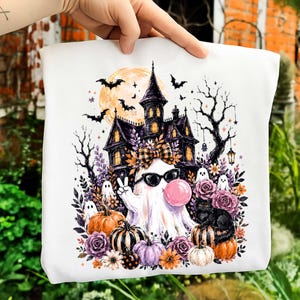 May include: A white sweatshirt featuring a graphic print of a ghost wearing sunglasses and blowing a pink bubble, positioned in front of a haunted house, a full moon, bats, pumpkins, purple roses, and a black cat with a plaid bow.