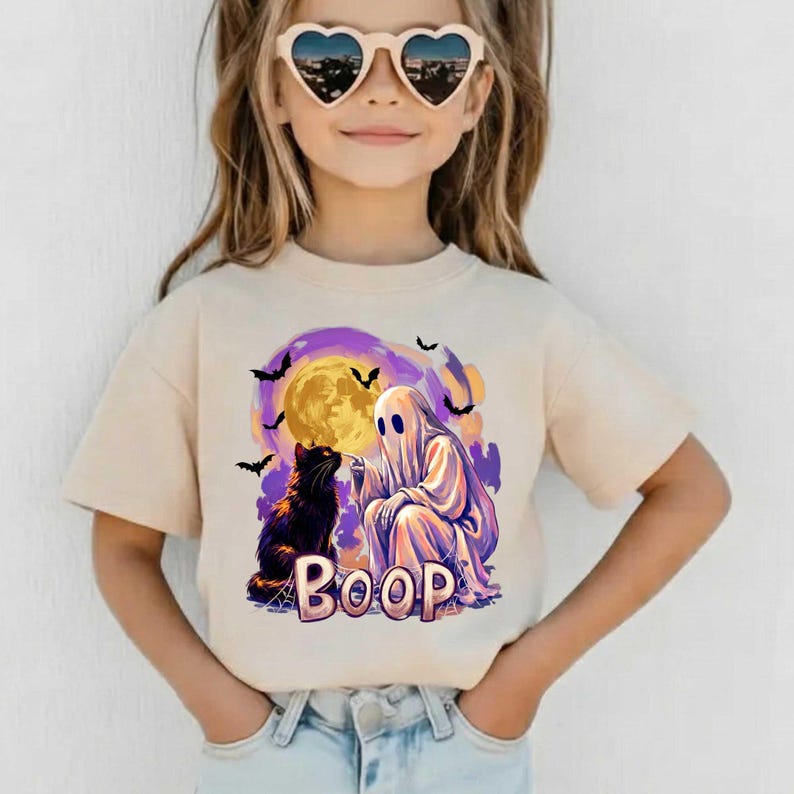 May include: A beige short-sleeve t-shirt featuring a graphic print of a white ghost touching the nose of a black cat. The design includes a large yellow moon, purple and orange clouds, black bats, and the word BOOP in large block letters.