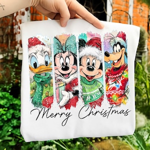 Puede incluir: Sudadera blanca con cuatro personajes de dibujos animados: Pato Donald, Minnie Mouse, Mickey Mouse y Goofy, todos vestidos para Navidad. Las palabras "Merry Christmas" están escritas debajo.