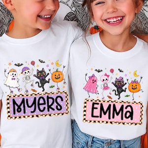May include: Two white t-shirts featuring Halloween graphics of a ghost, skeleton, black cat, and pumpkin. The left shirt displays the name MYERS in a purple box, and the right shirt displays the name EMMA in a pink box with a checkered border.