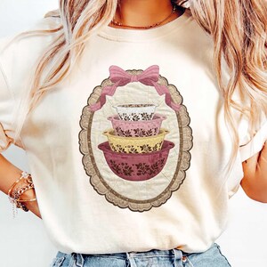 May include: Cream-colored t-shirt featuring a decorative oval design with a stack of four floral-patterned mixing bowls in shades of pink, yellow, and white. A pink bow adorns the top of the oval, adding a touch of elegance.